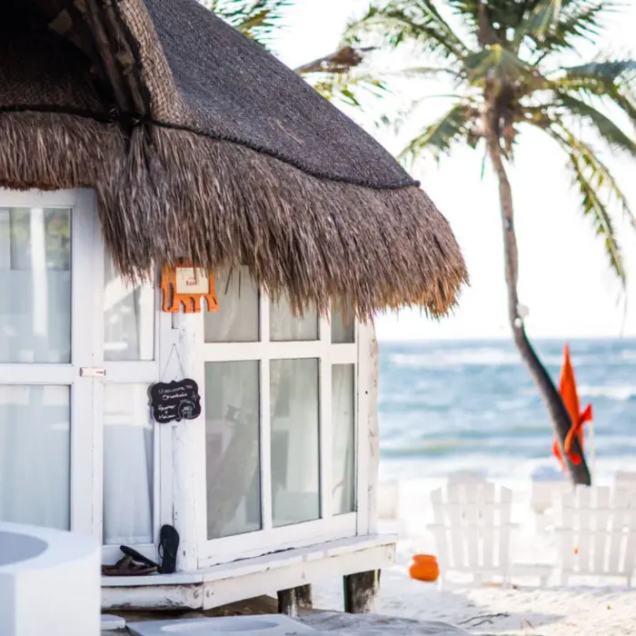 Close-up exterior of a beach casita with a thatched roof, white walls, and view of the beach and ocean.