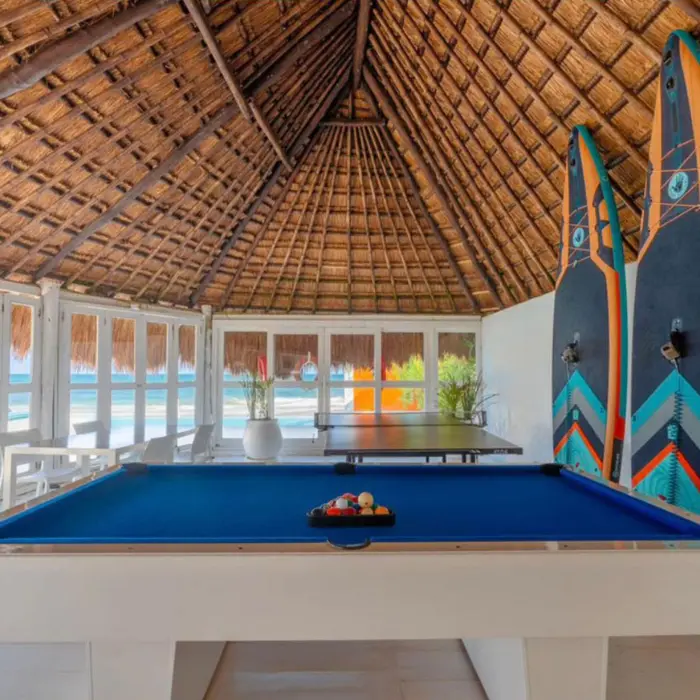 Thatched-roof game area with a blue pool table, table tennis, and paddleboards, overlooking the beach.