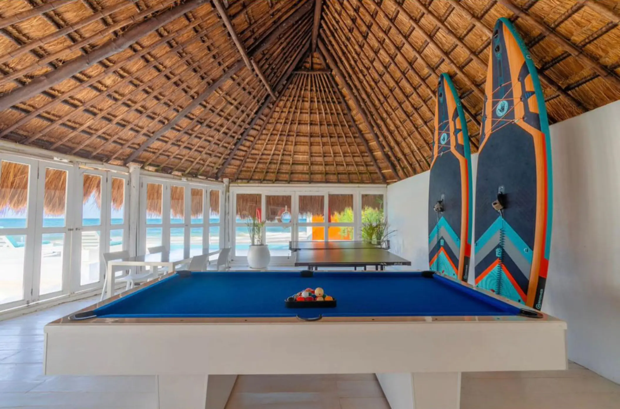 Thatched-roof game area with a blue pool table, table tennis, and paddleboards, overlooking the beach.