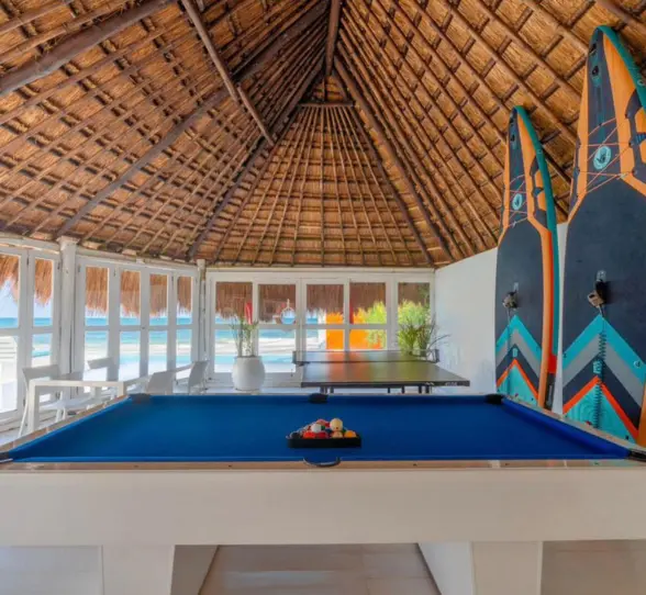 Thatched-roof game area with a blue pool table, table tennis, and paddleboards, overlooking the beach.