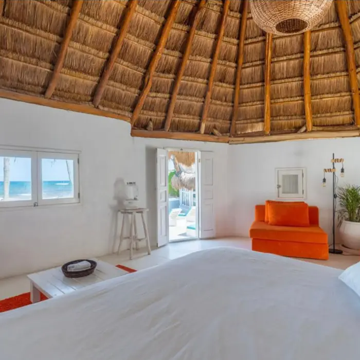 Interior of a thatched-roof casita with vibrant orange seating, a large bed, and ocean views through windows.