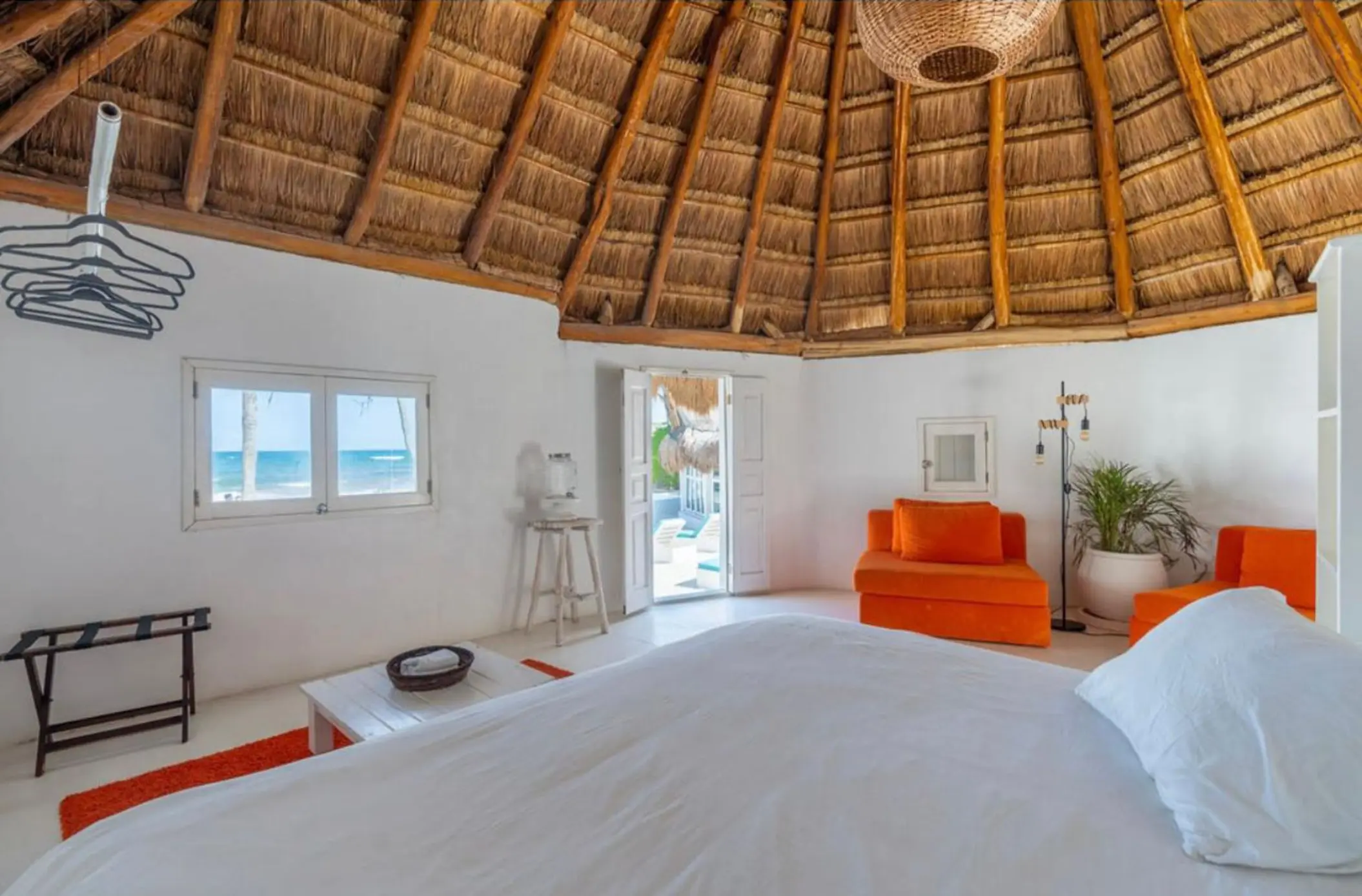 Interior of a thatched-roof casita with vibrant orange seating, a large bed, and ocean views through windows.