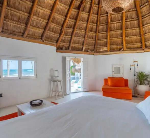 Interior of a thatched-roof casita with vibrant orange seating, a large bed, and ocean views through windows.
