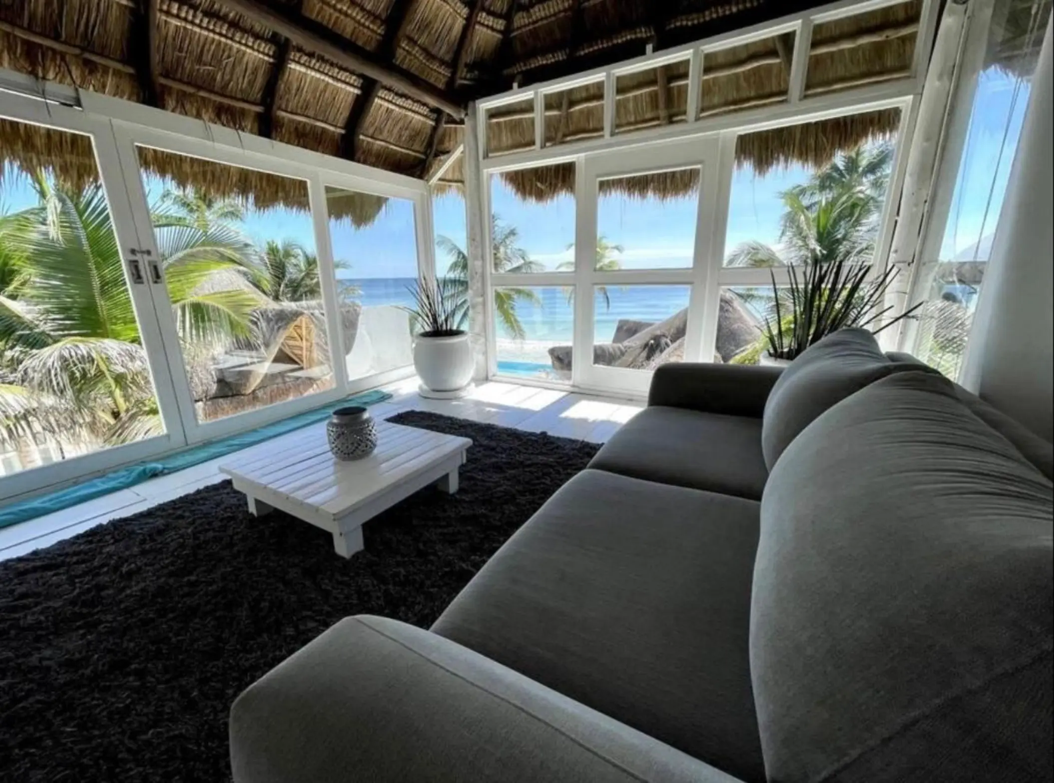 A living area with gray seating, a thatched roof, and floor-to-ceiling windows providing panoramic ocean views.
