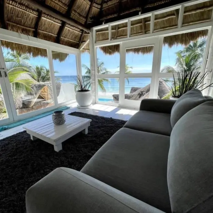 A living area with gray seating, a thatched roof, and floor-to-ceiling windows providing panoramic ocean views.
