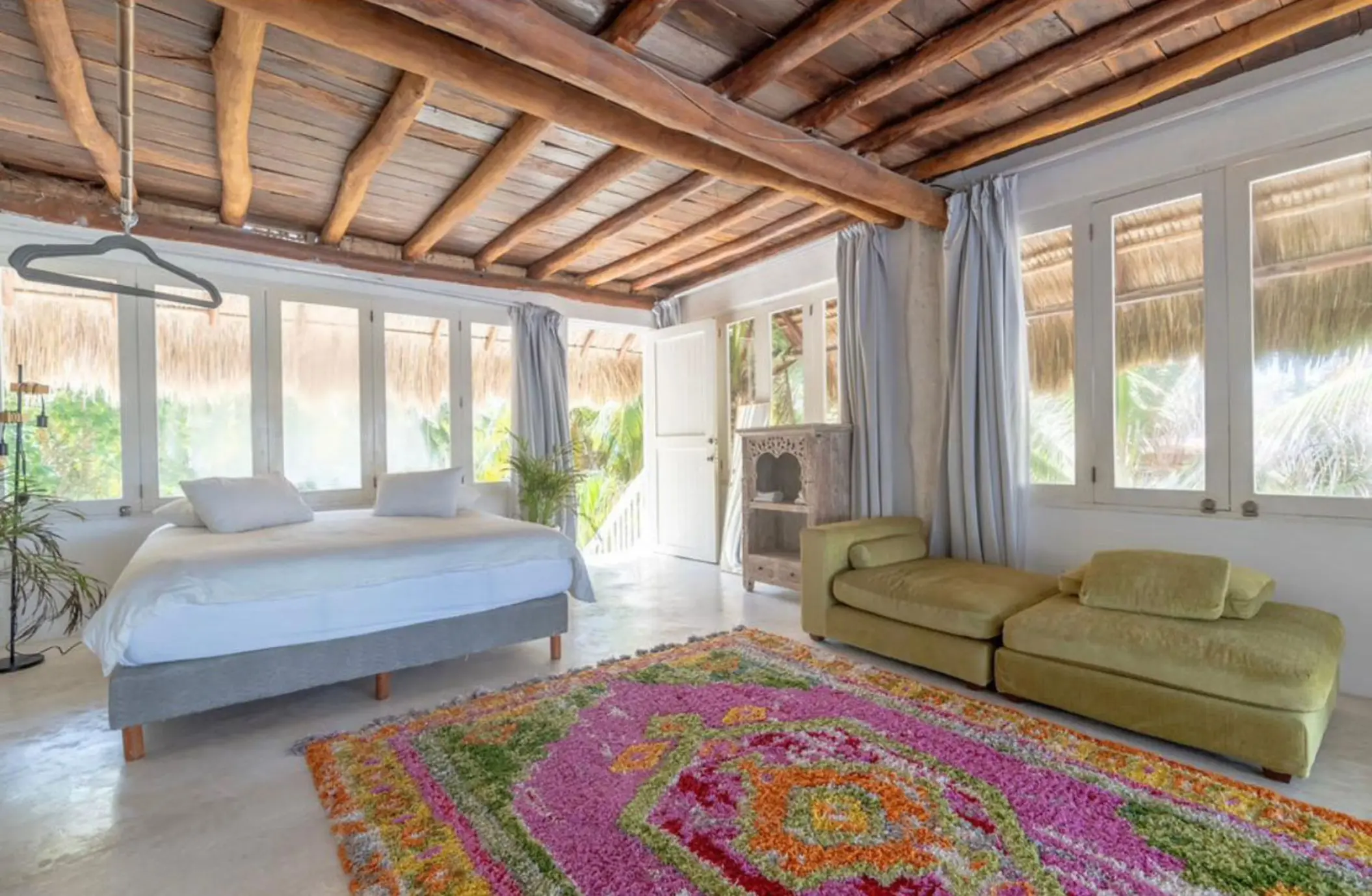 A penthouse suite with a wooden beamed ceiling, a large bed, green seating, and windows overlooking the surrounding foliage.