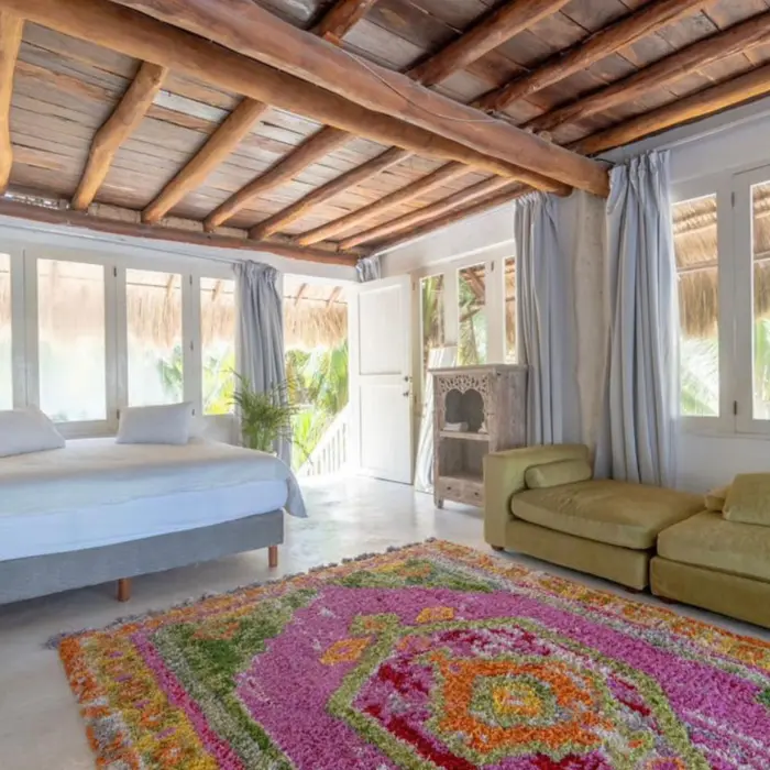 A penthouse suite with a wooden beamed ceiling, a large bed, green seating, and windows overlooking the surrounding foliage.