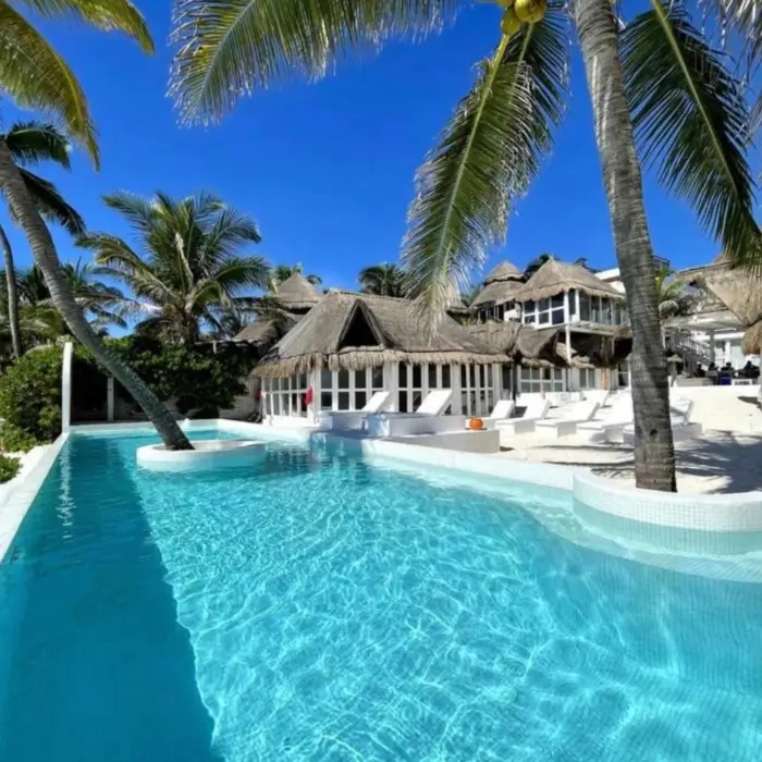 Bright turquoise pool surrounded by palm trees, white lounge chairs, and thatched-roof buildings.