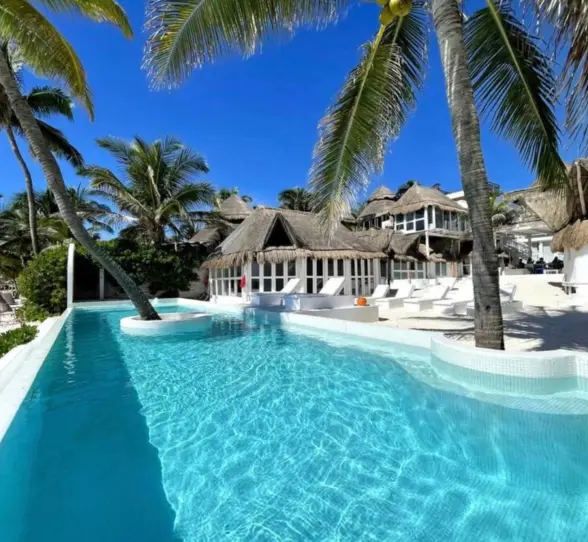Bright turquoise pool surrounded by palm trees, white lounge chairs, and thatched-roof buildings.