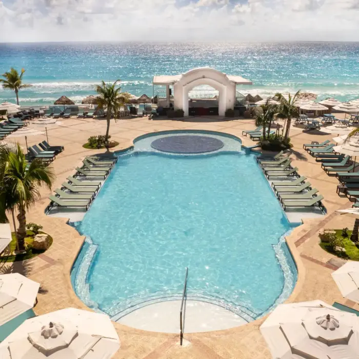An aerial view shows the large pool area, lounge chairs, and palm trees next to a white sand beach.