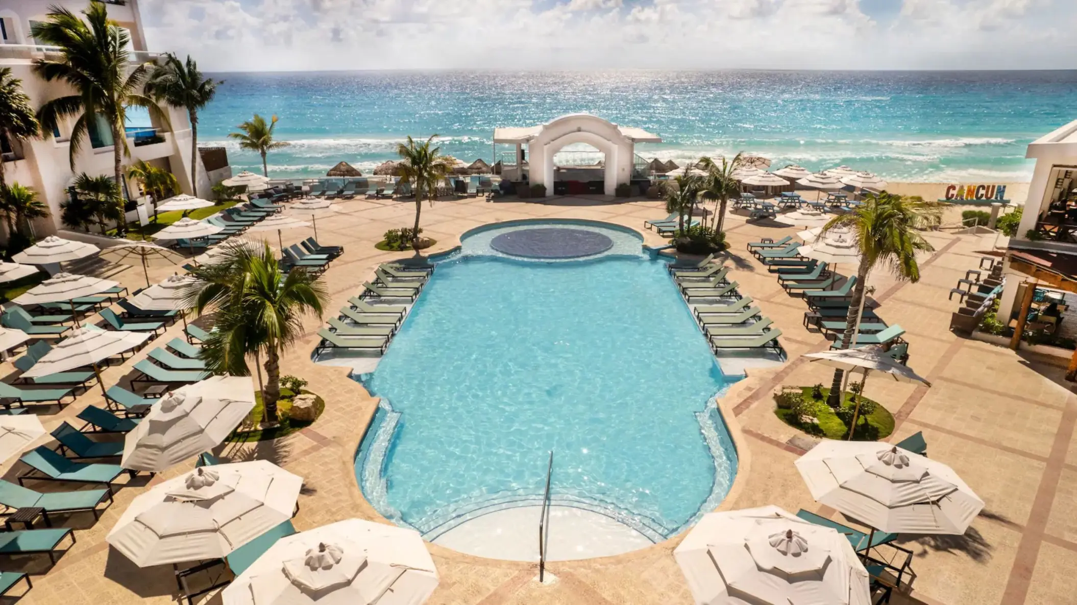 An aerial view shows the large pool area, lounge chairs, and palm trees next to a white sand beach.