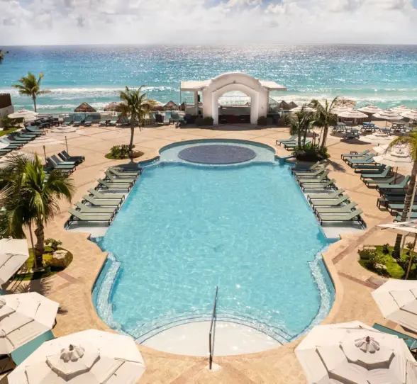 An aerial view shows the large pool area, lounge chairs, and palm trees next to a white sand beach.