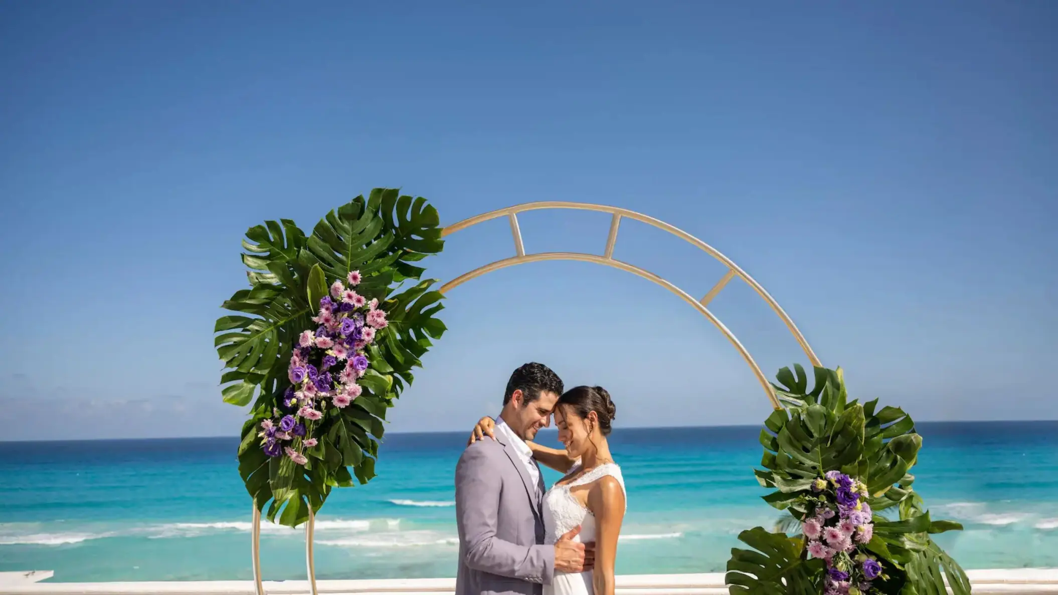 A newlywed couple embraces under a floral archway with the vibrant blue ocean in the background.