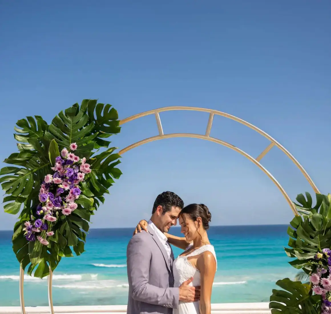 A newlywed couple embraces under a floral archway with the vibrant blue ocean in the background.