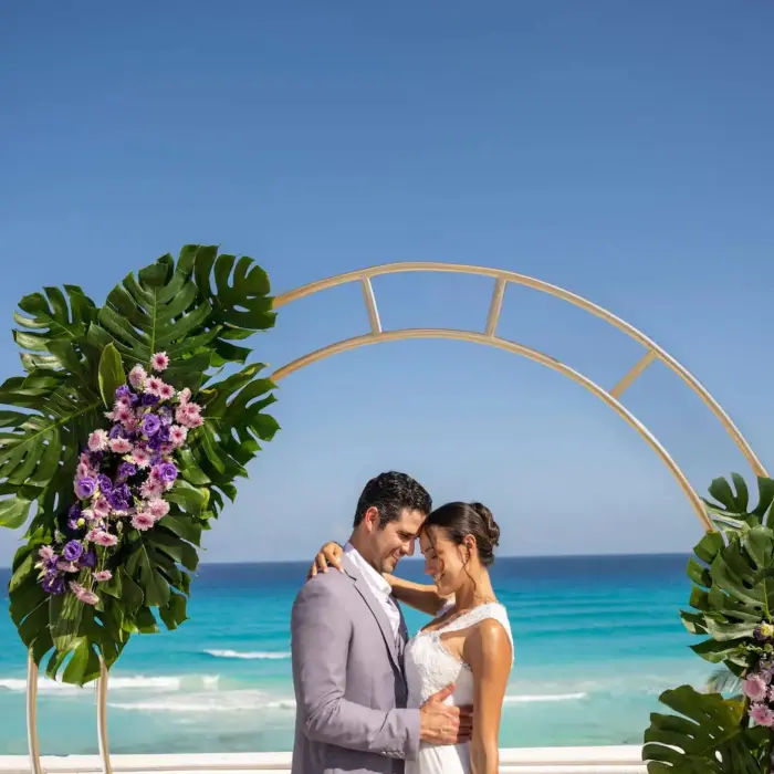A newlywed couple embraces under a floral archway with the vibrant blue ocean in the background.