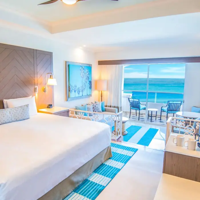 A bright hotel room with a large bed, a sofa, and a balcony with an oceanfront view.