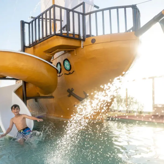 A child is seen splashing at the bottom of a waterslide from a large pirate ship in the kids pool.