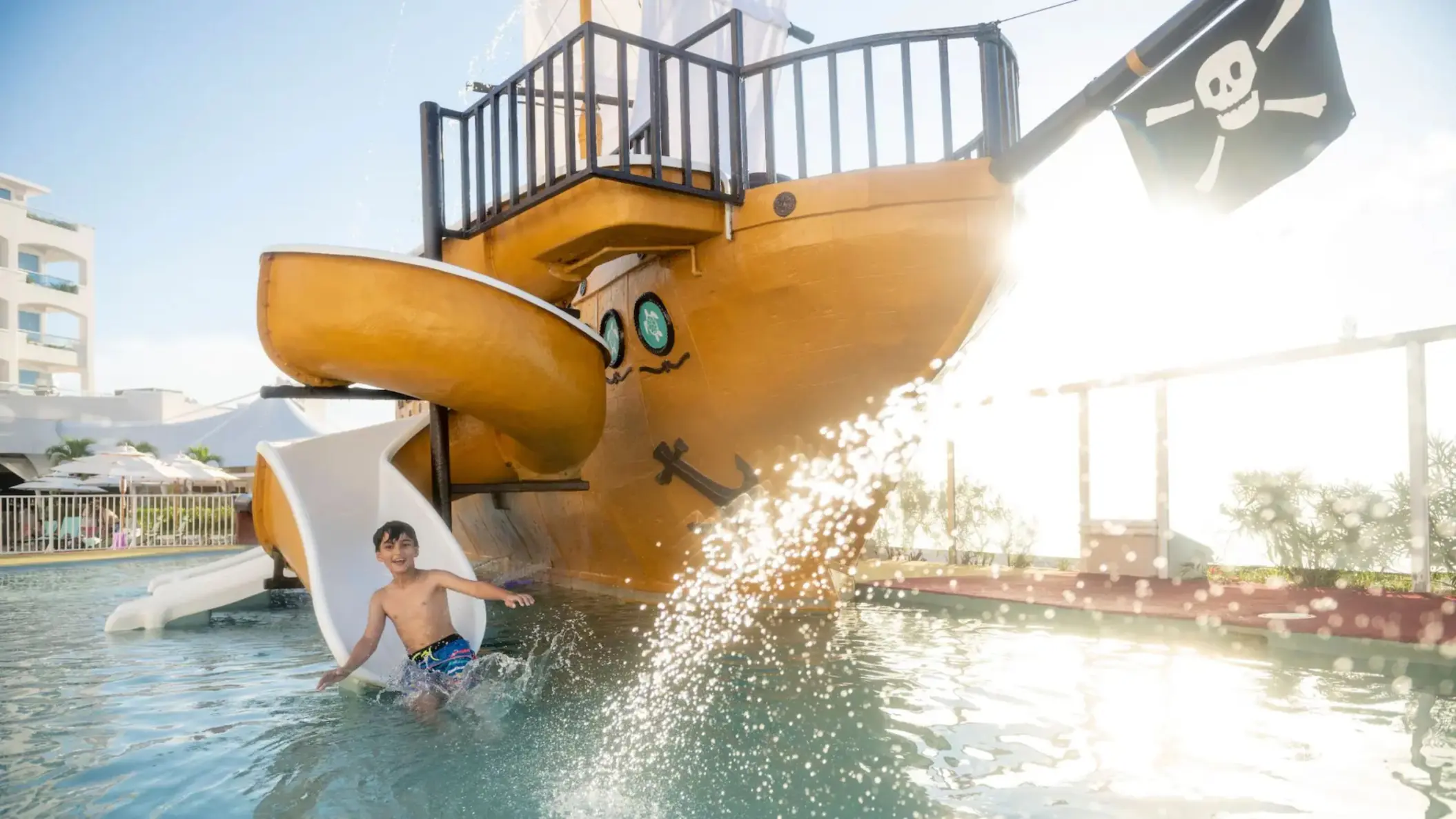 A child is seen splashing at the bottom of a waterslide from a large pirate ship in the kids pool.