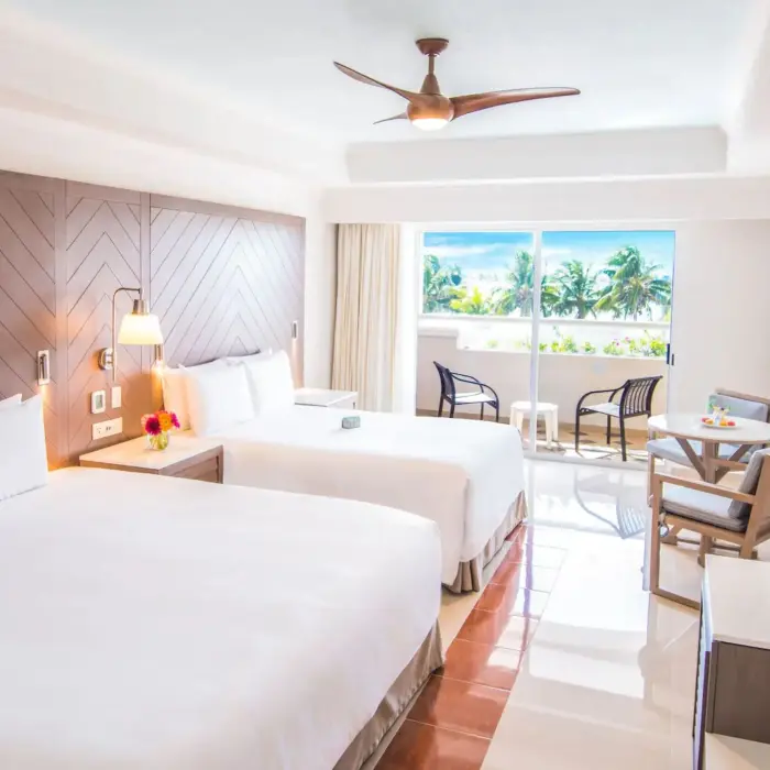 A hotel room with two large beds, a table, and a balcony overlooking the palm trees and the sea.