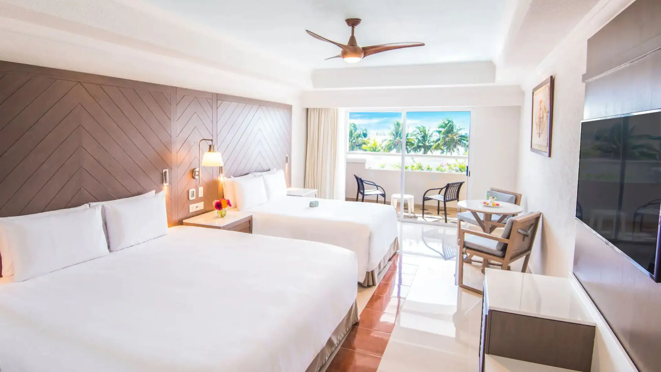 A hotel room with two large beds, a table, and a balcony overlooking the palm trees and the sea.