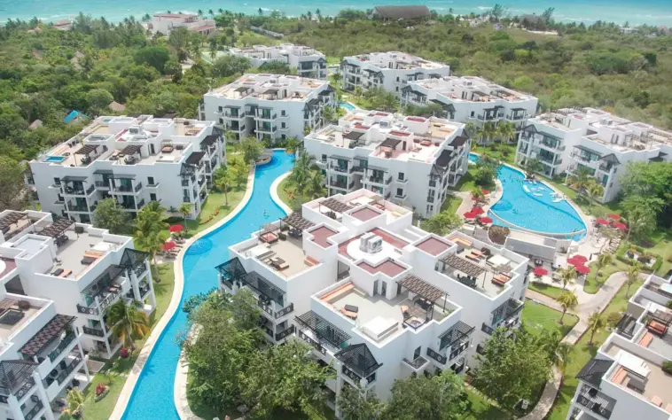 Aerial view of The Fives Beach Hotel, showcasing white resort buildings with terraces, surrounded by a long, winding river-style pool and green vegetation, leading towards the ocean.