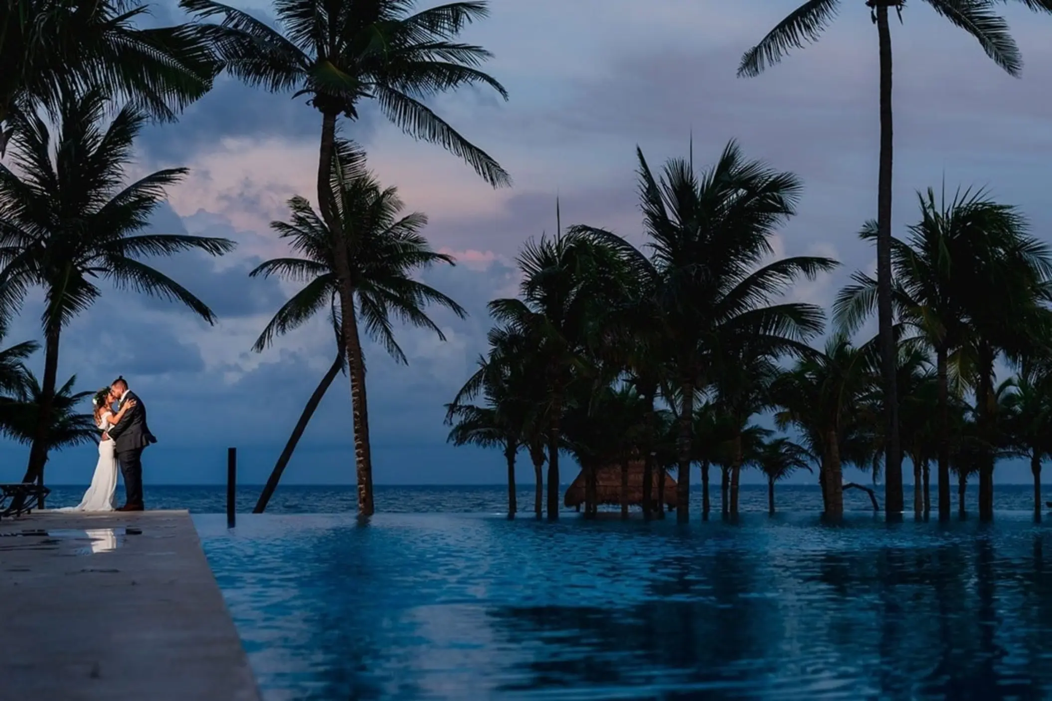 A romantic view of a bride and groom embracing at dusk by an infinity pool overlooking the ocean with palm trees silhouetted against the colorful sky.