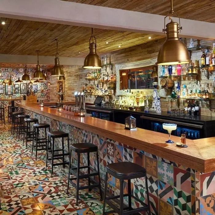 A vibrant bar scene at Cantina 1910 with a long wooden bar counter, rustic lighting, and patterned floor tiles, showcasing various bottles and stools.