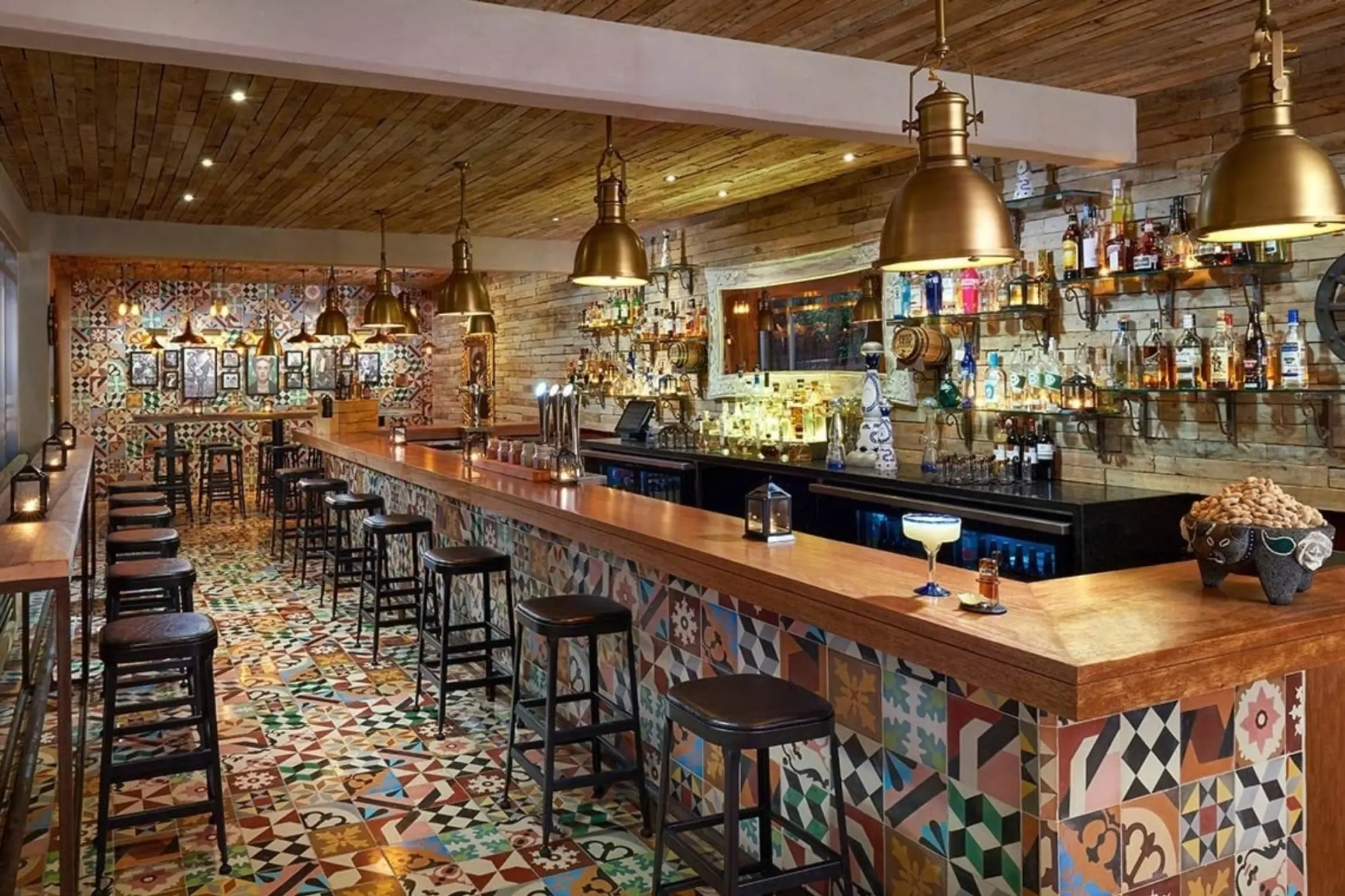 A vibrant bar scene at Cantina 1910 with a long wooden bar counter, rustic lighting, and patterned floor tiles, showcasing various bottles and stools.
