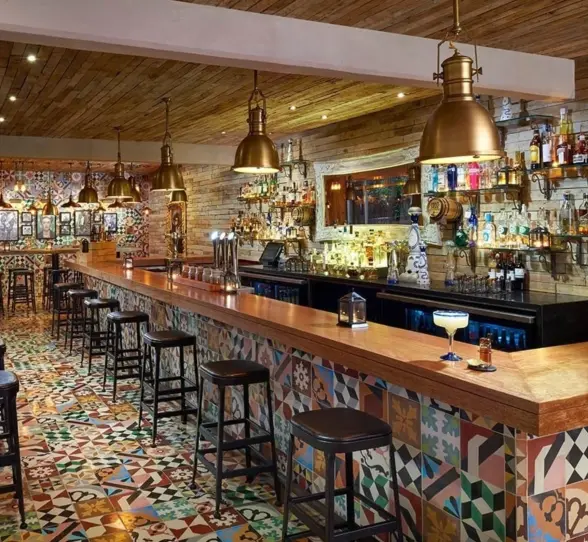 A vibrant bar scene at Cantina 1910 with a long wooden bar counter, rustic lighting, and patterned floor tiles, showcasing various bottles and stools.