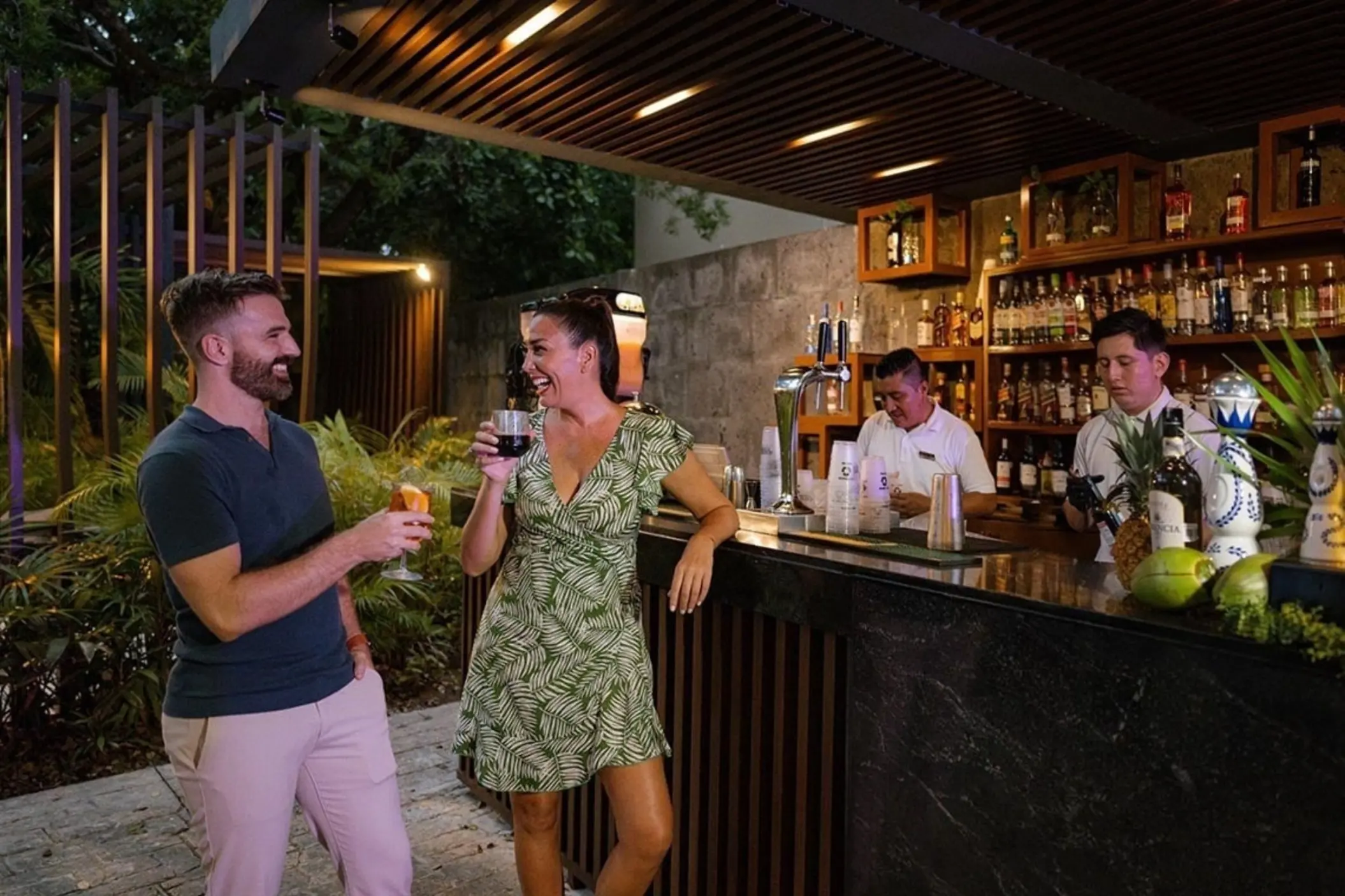 A couple smiling and enjoying drinks at a stylish outdoor bar, with bartenders preparing cocktails in the background.