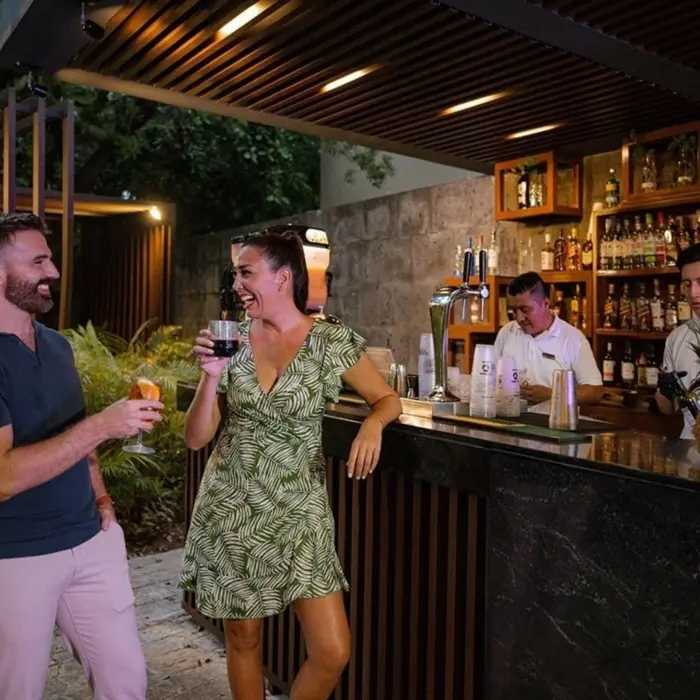 A couple smiling and enjoying drinks at a stylish outdoor bar, with bartenders preparing cocktails in the background.