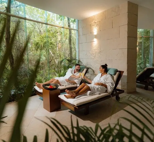 A couple relaxing on lounge chairs at the spa, wrapped in white robes, enjoying drinks in a tranquil setting with natural light and lush greenery.