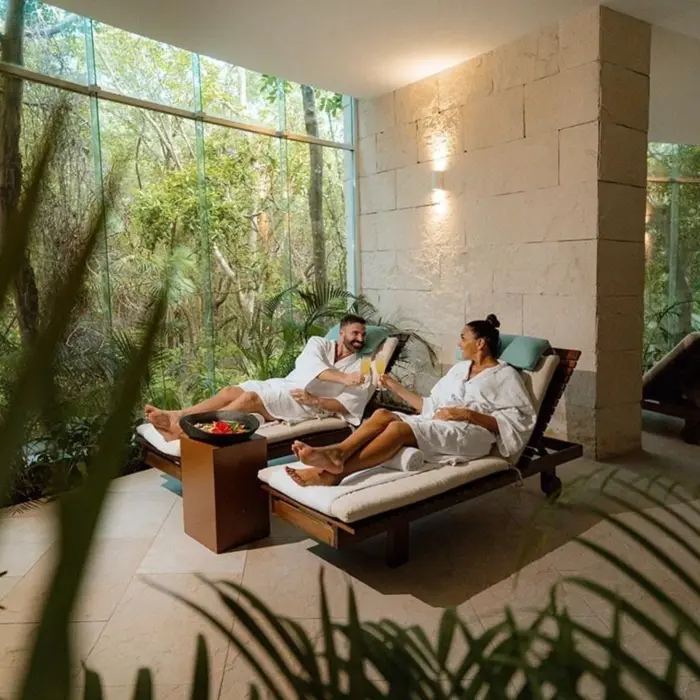 A couple relaxing on lounge chairs at the spa, wrapped in white robes, enjoying drinks in a tranquil setting with natural light and lush greenery.