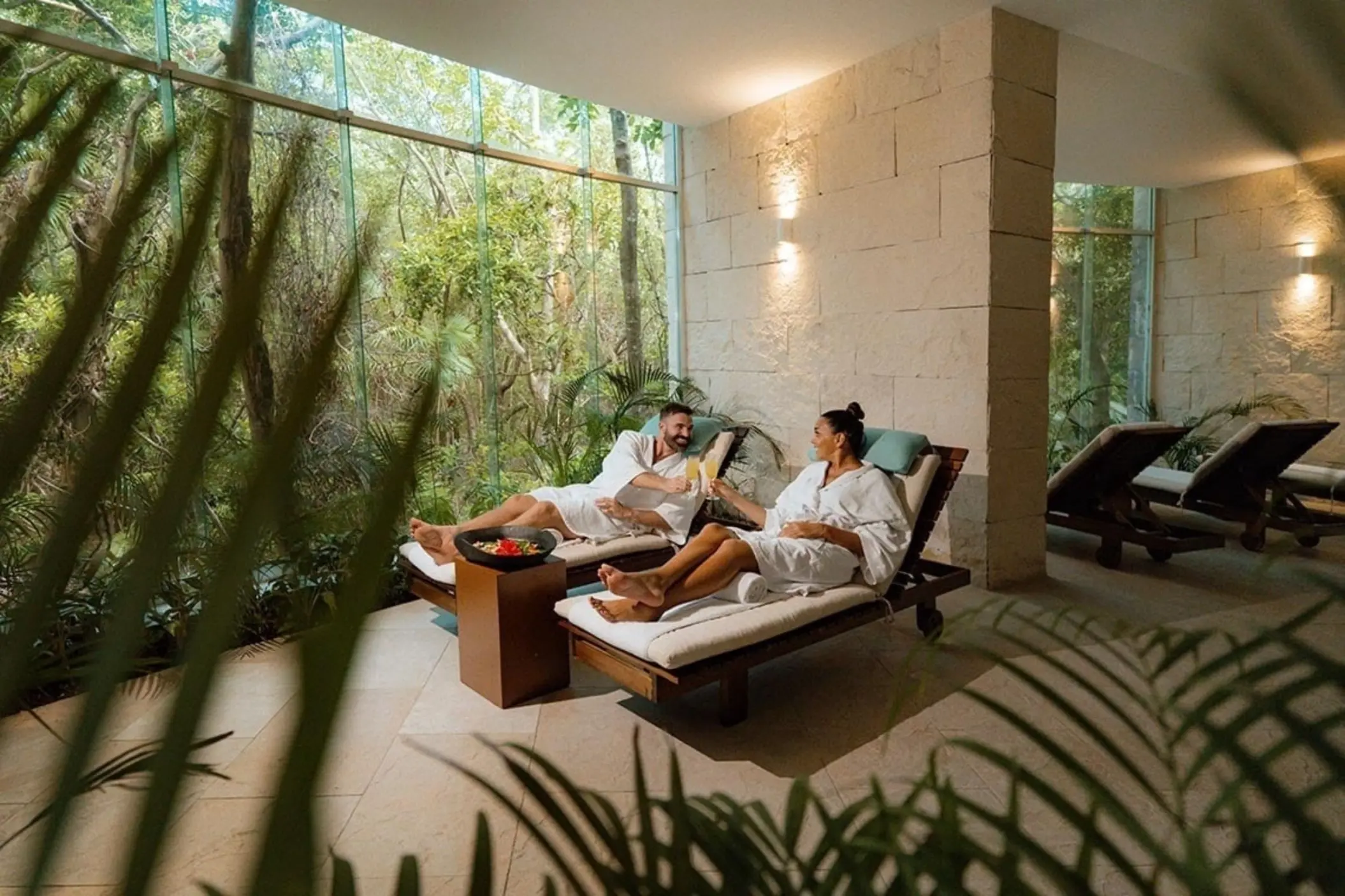 A couple relaxing on lounge chairs at the spa, wrapped in white robes, enjoying drinks in a tranquil setting with natural light and lush greenery.