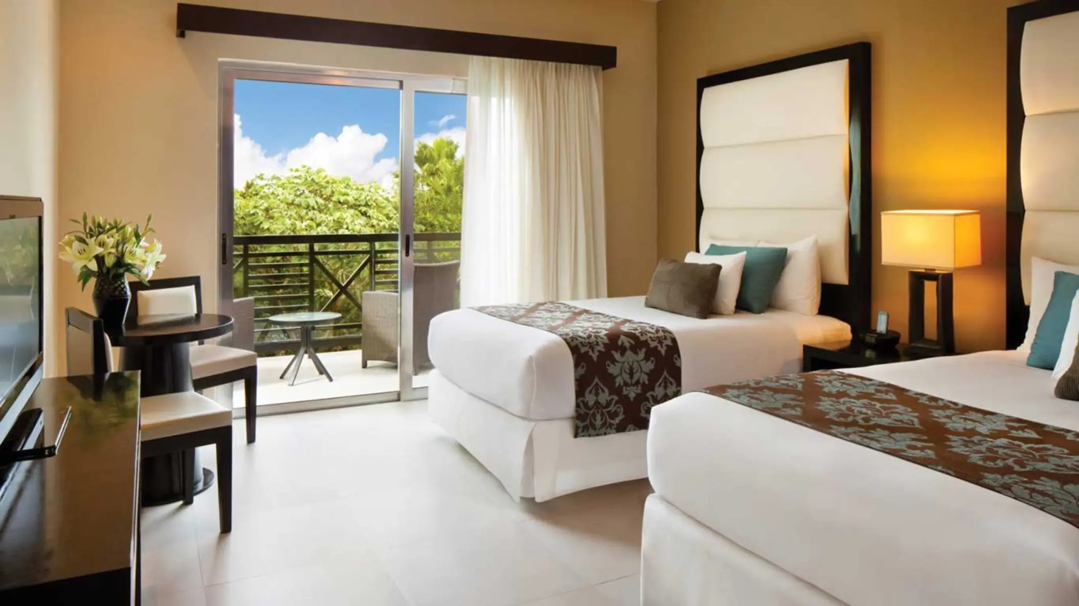 A bright and spacious deluxe suite with two comfortable beds, a sitting area, and a balcony offering a view of the surrounding greenery.