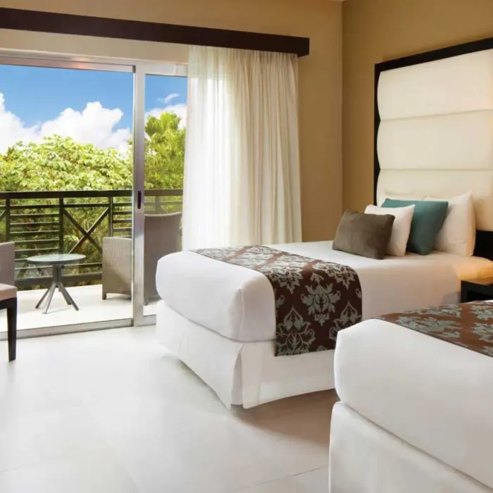 A bright and spacious deluxe suite with two comfortable beds, a sitting area, and a balcony offering a view of the surrounding greenery.