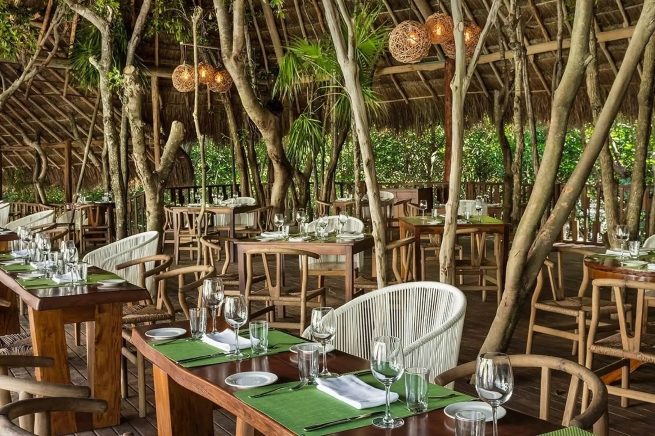 An inviting outdoor dining area at Natura restaurant, featuring wooden tables, wicker chairs, and rustic lighting fixtures nestled among lush trees and thatched roofs.