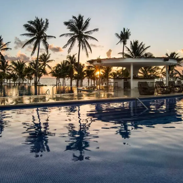 A stunning infinity pool view at sunset, featuring a swim-up bar surrounded by palm trees and reflections of the vibrant sky on the water.