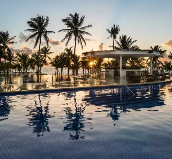 A stunning infinity pool view at sunset, featuring a swim-up bar surrounded by palm trees and reflections of the vibrant sky on the water.