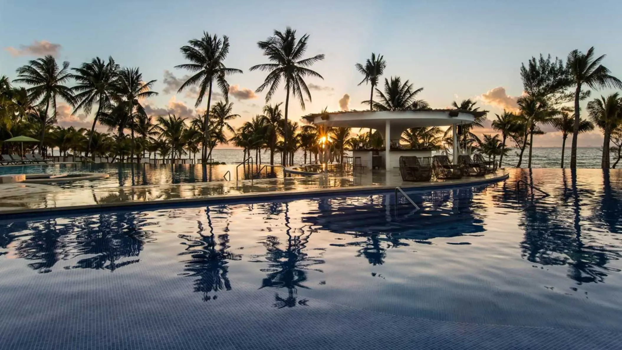 A stunning infinity pool view at sunset, featuring a swim-up bar surrounded by palm trees and reflections of the vibrant sky on the water.