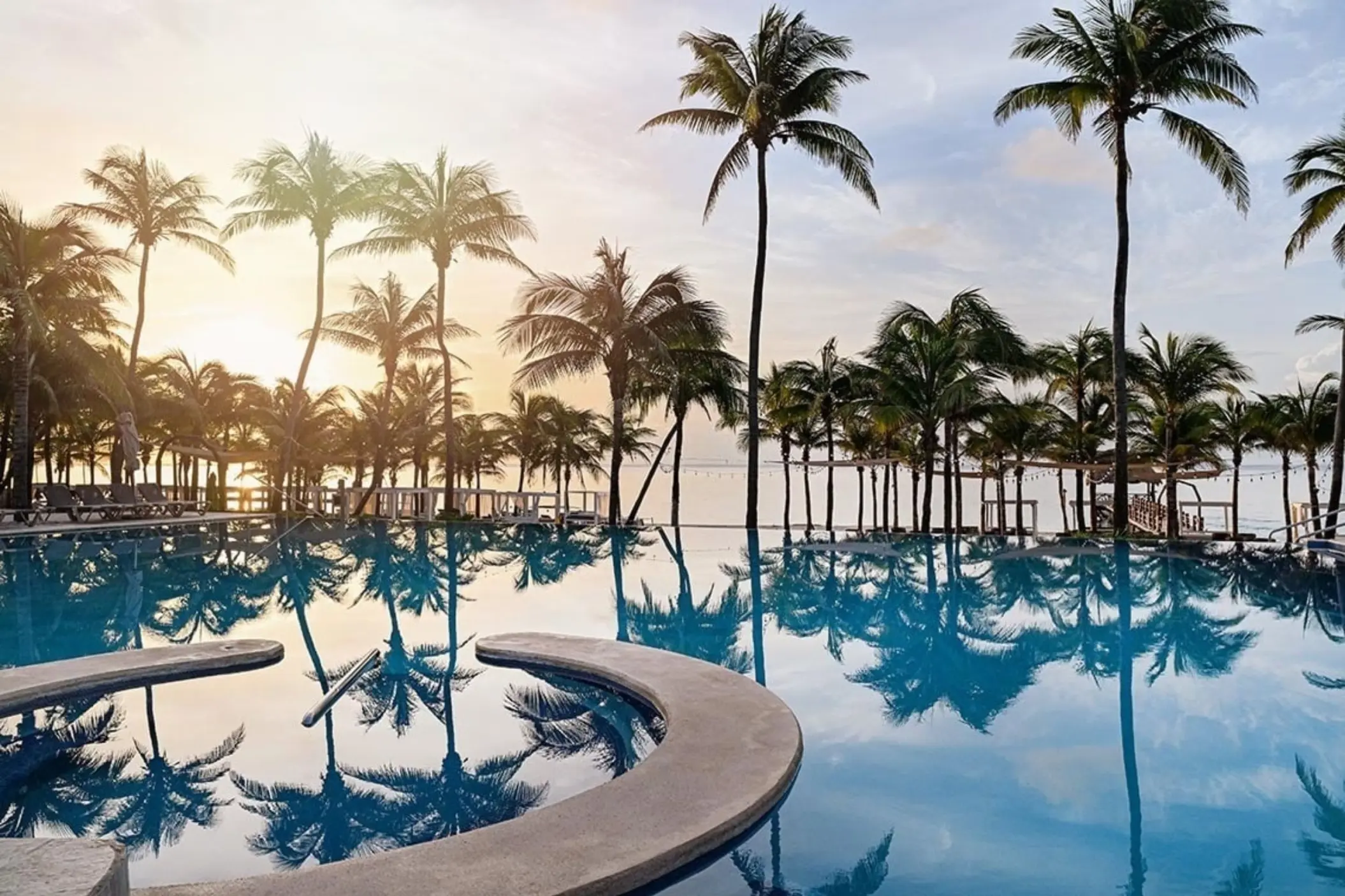 A panoramic view of a large swimming pool surrounded by numerous palm trees, with reflections on the calm water and the sky visible in the distance.