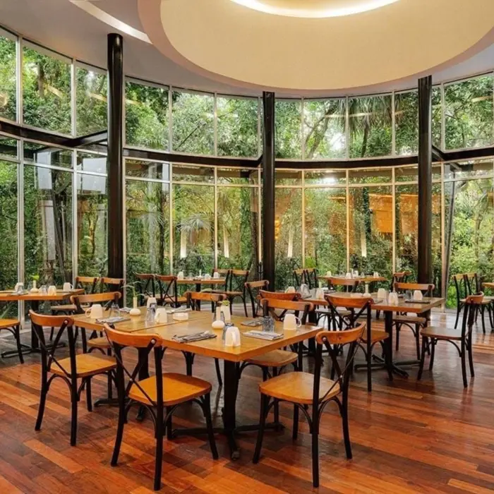 A bright and airy restaurant with floor-to-ceiling windows overlooking lush greenery, featuring modern wooden tables and chairs, and warm lighting.
