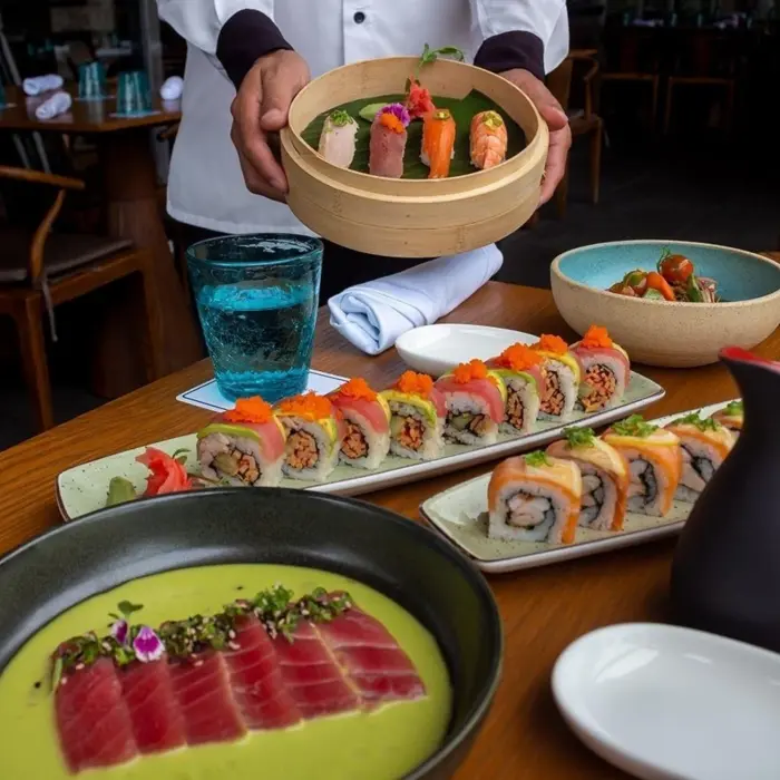 A professional chef presenting a platter of beautifully arranged sushi and sashimi, with various dishes, including tuna slices and sushi rolls, on a wooden table.