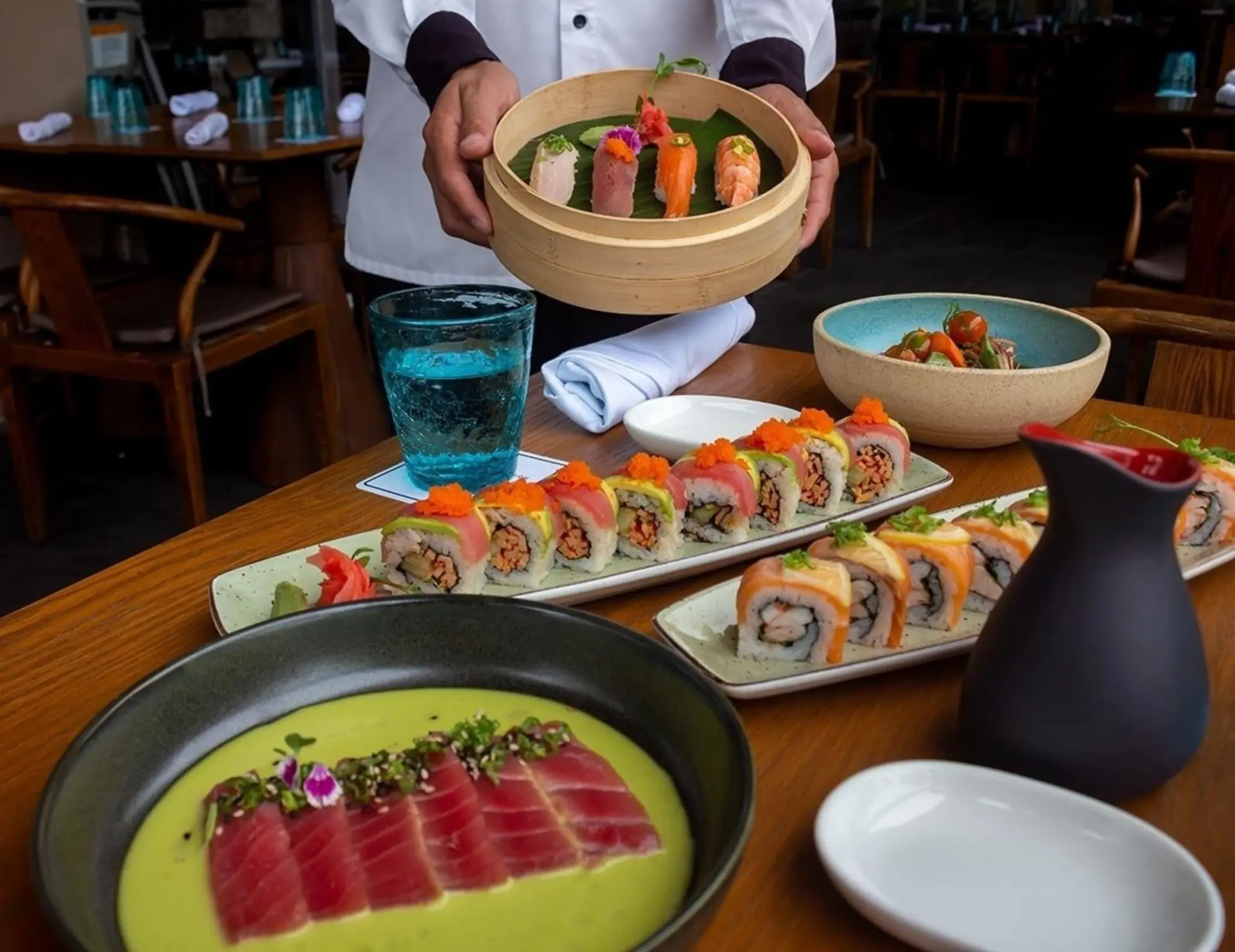 A professional chef presenting a platter of beautifully arranged sushi and sashimi, with various dishes, including tuna slices and sushi rolls, on a wooden table.
