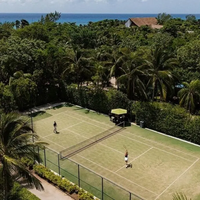 An aerial view of a tennis court surrounded by dense greenery and palm trees, with players engaged in a match, and the ocean visible in the background.