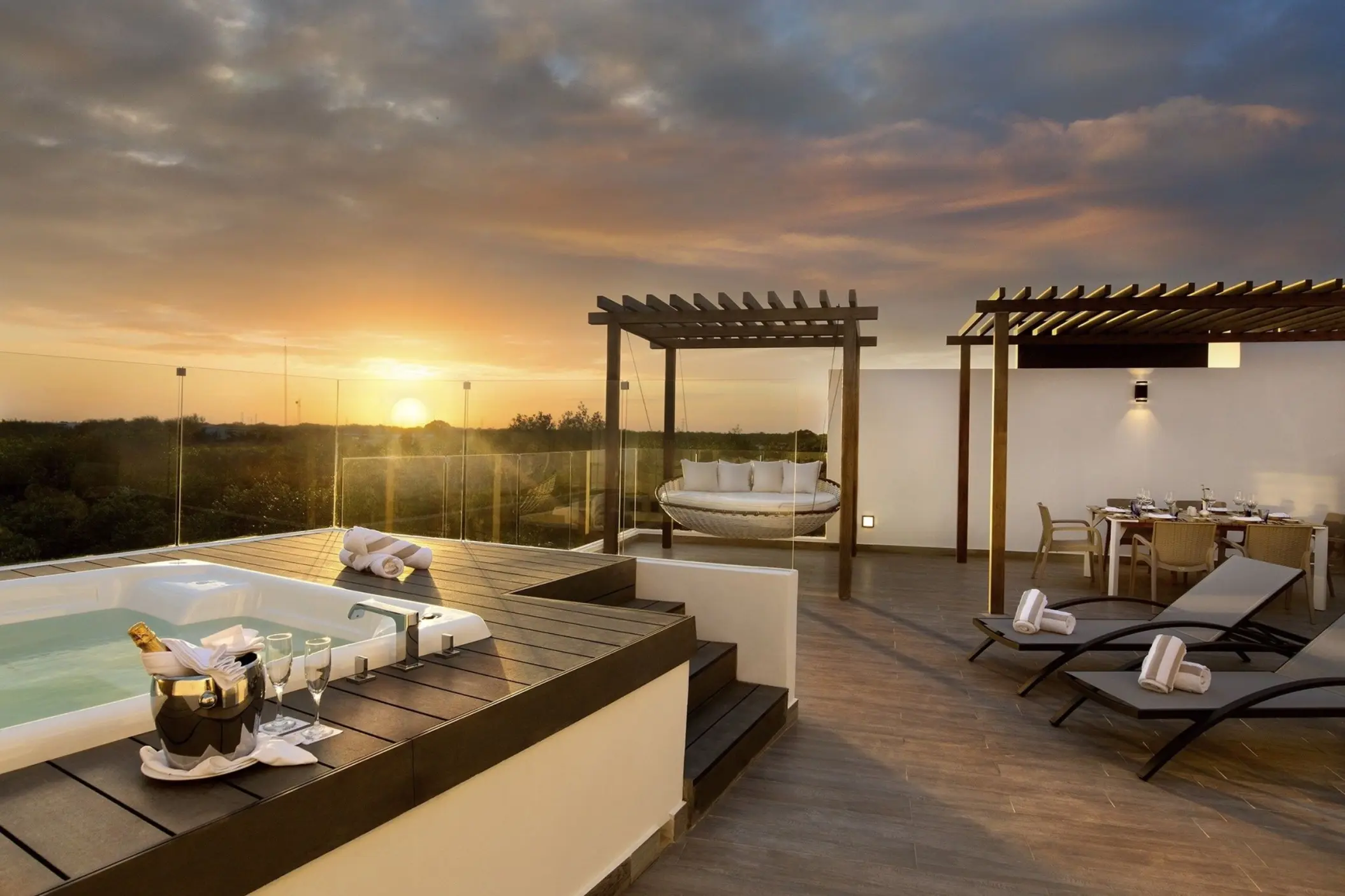 A luxurious penthouse terrace at sunset, featuring a hot tub, comfortable lounge chairs, a covered dining area, and a large daybed swing.