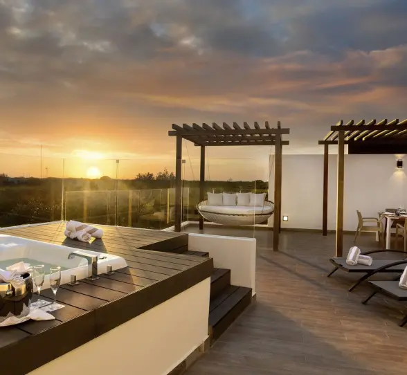 A luxurious penthouse terrace at sunset, featuring a hot tub, comfortable lounge chairs, a covered dining area, and a large daybed swing.