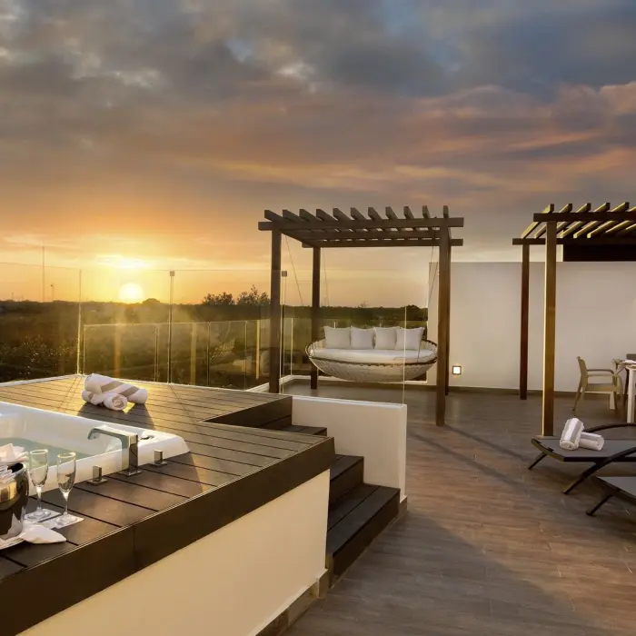 A luxurious penthouse terrace at sunset, featuring a hot tub, comfortable lounge chairs, a covered dining area, and a large daybed swing.