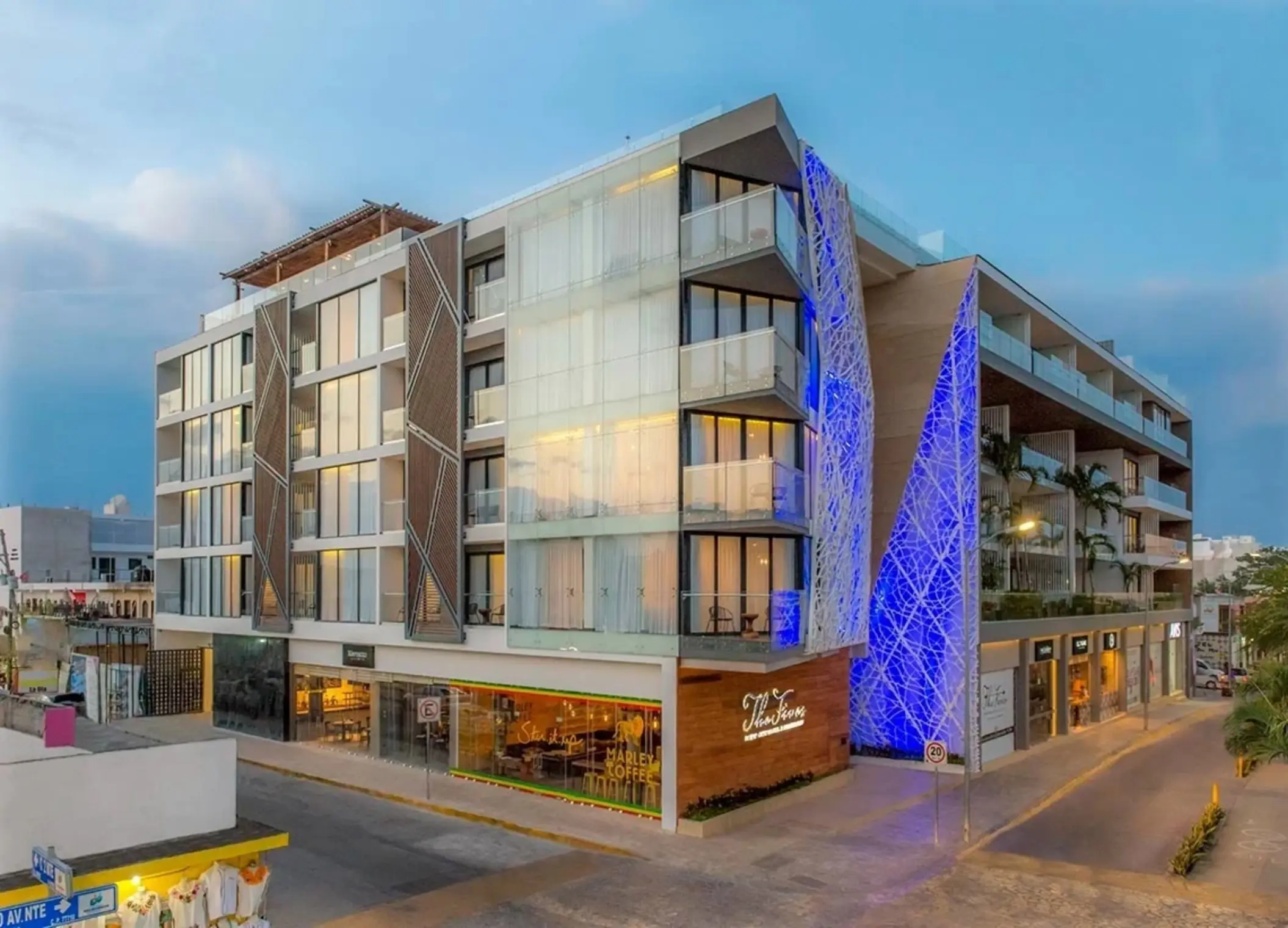 Exterior view of The Fives Downtown Hotel lit up in blue with visible balconies and Marley Coffee at street level.