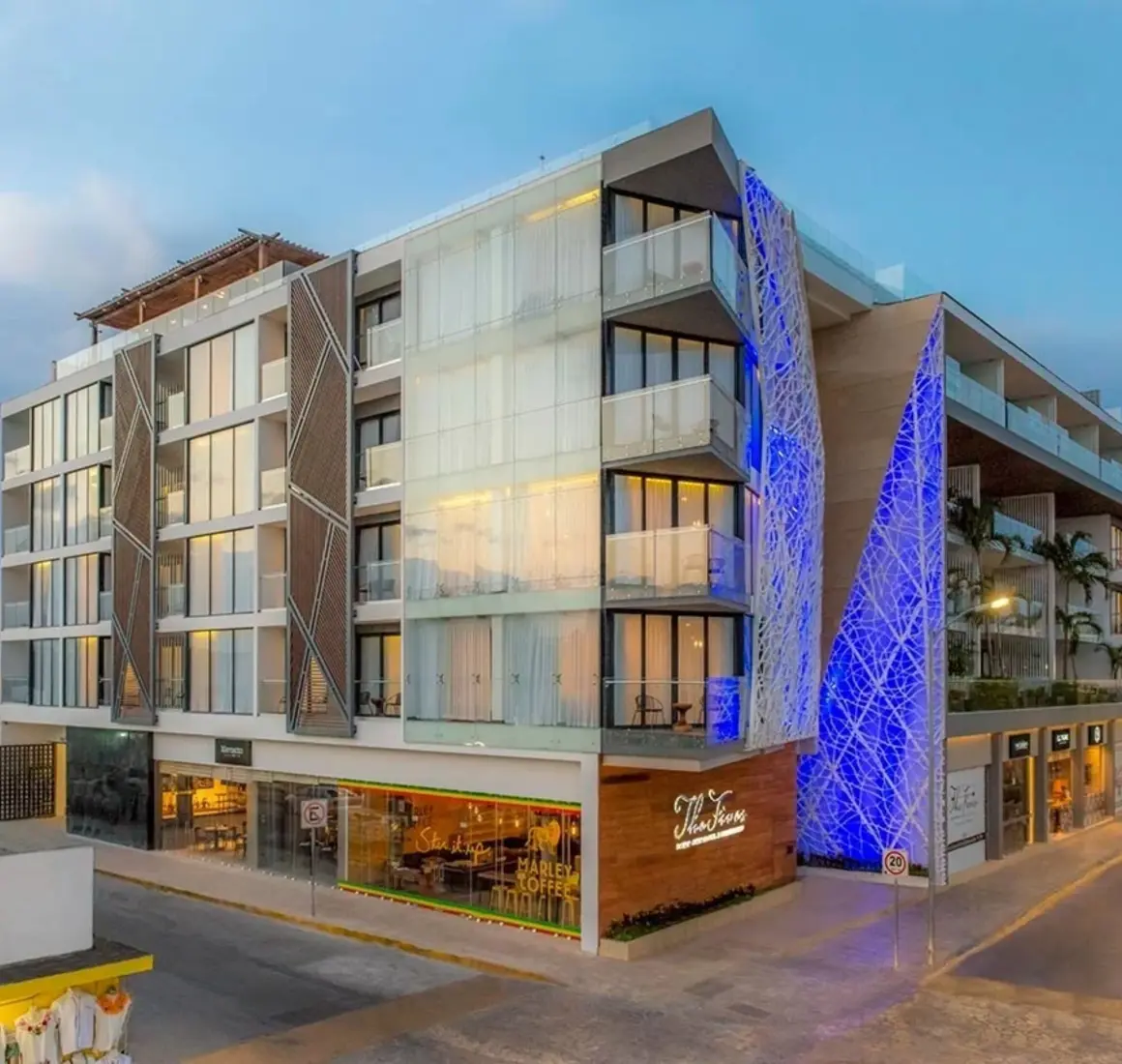 Exterior view of The Fives Downtown Hotel lit up in blue with visible balconies and Marley Coffee at street level.